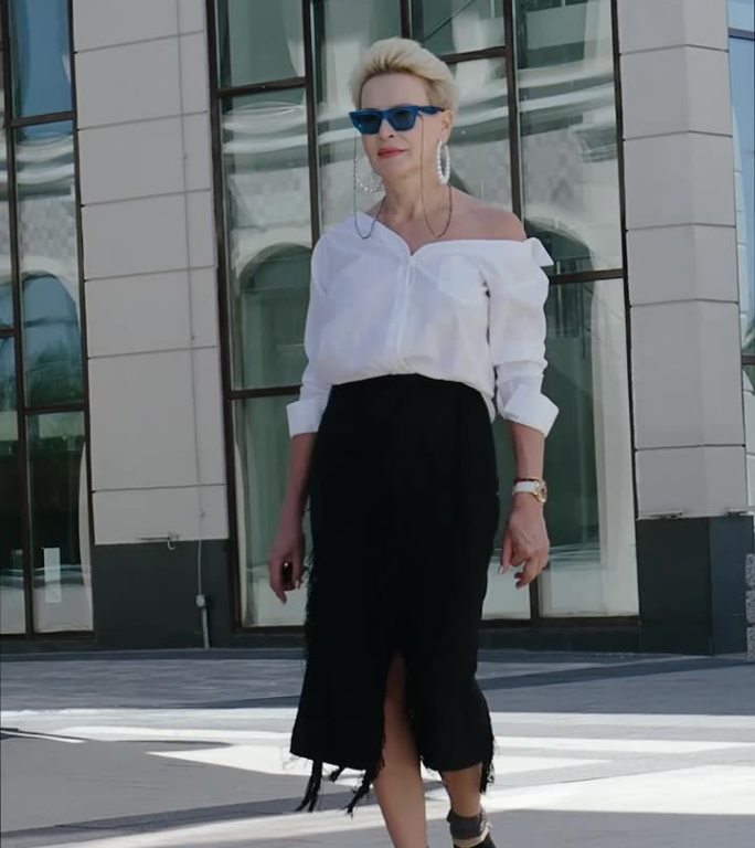 自信和成熟的成熟女人走在城市的街道上，时尚的整体别致的白衬衫和黑色流苏裙搭配高跟凉鞋和袜子，时尚的风