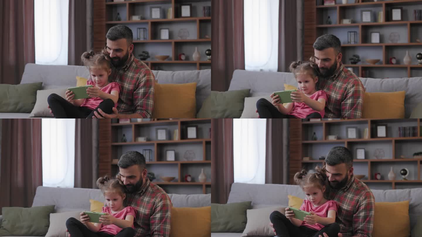 阖家欢乐。年轻的父亲和可爱的女儿在智能手机上看有趣的视频或玩游戏。爸爸带着小女儿尽情地共度时光，在家