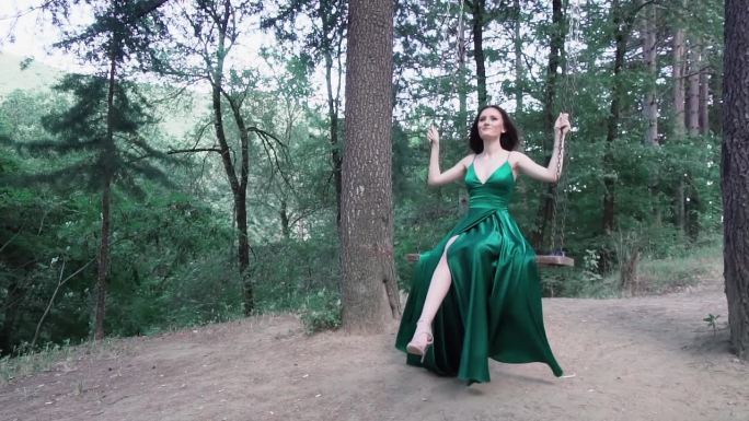 年轻美丽的女人坐在秋千上，穿着豪华的绿色丝绸连衣裙，拖着长长的火车，在皇家公园摆姿势。拍摄优雅的舞会
