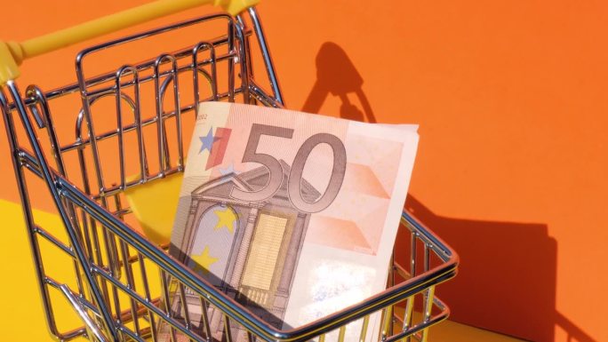 手加50欧元钞票在玩具超市购物车黄色背景的钱。销售一篮子欧元票据。最低生活工资概念:贷款、投资养老、