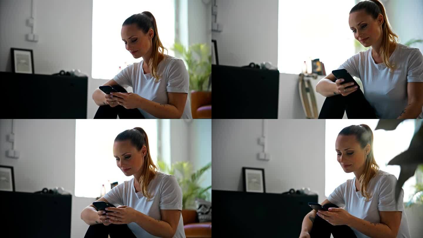 微笑的年轻女子扎着马尾辫坐在家里客厅的地板上用智能手机发短信。生活方式，居家生活，简单生活。以8K分
