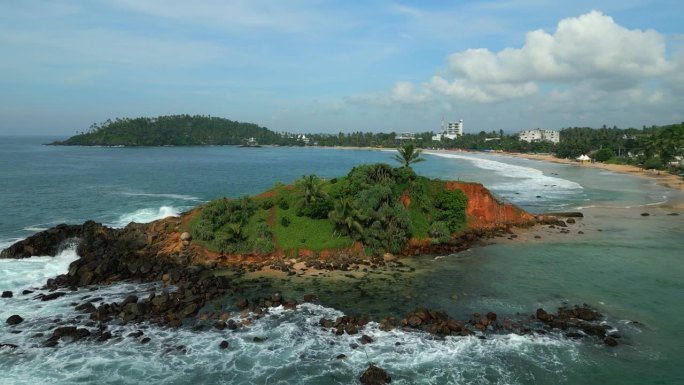 绿色的热带岩石岛在蓝绿色的海洋中间被海浪冲刷垂直鸟瞰。无人机拍摄的画面显示，斯里兰卡米里萨的鹦鹉岩被