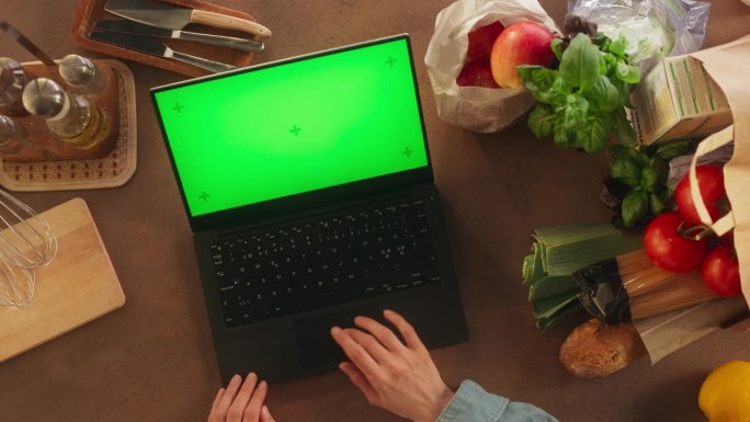顶部静态视图匿名妇女使用笔记本电脑与模拟绿色屏幕Chromakey显示在厨房桌子上。女厨师写博客，分