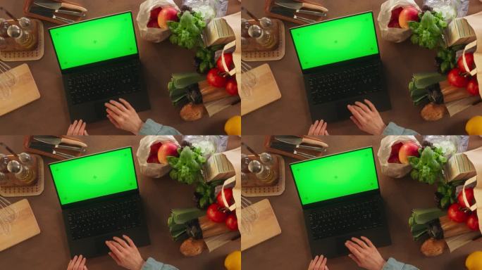 顶部静态视图匿名妇女使用笔记本电脑与模拟绿色屏幕Chromakey显示在厨房桌子上。女厨师写博客，分