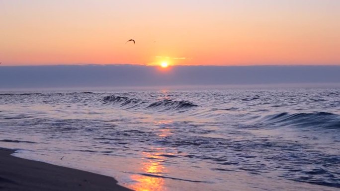 海边美丽的日出。太阳从地平线上的天空云后升起，在蓝色的大海上，海浪在沙滩上翻滚。美丽的海景海景。自然