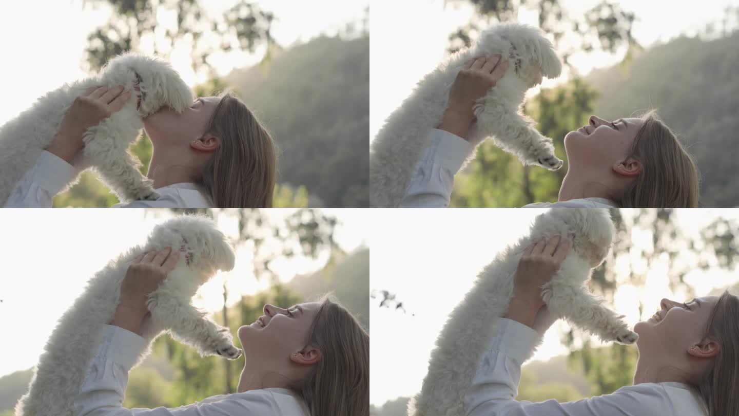 一个女孩和她的白狗的吻很小。那个女人把她怀里的狗举到她面前，又踢又摸。晴朗的夏日。户外休闲