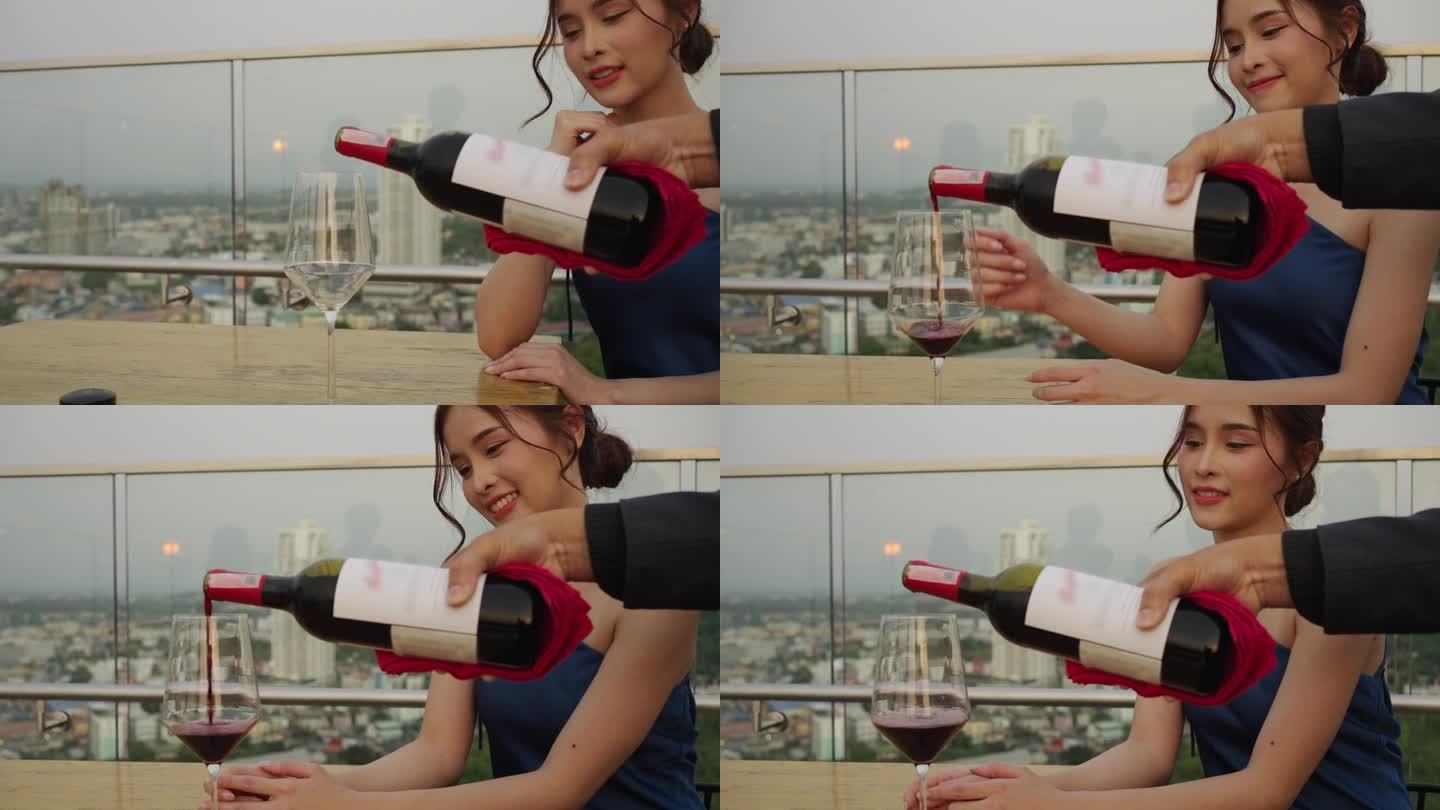 在一家城市酒店的屋顶酒吧里，男服务员拿着一瓶红酒，为一位美丽的亚洲女孩斟酒。