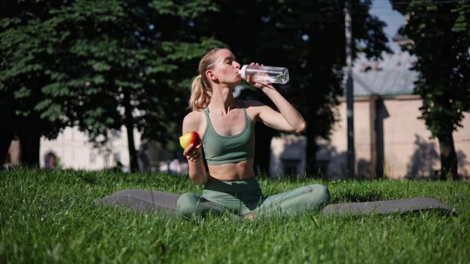 运动女孩坐成莲花式，从运动瓶里喝水，手里拿着苹果，强调健康，适当的饮食和积极的生活方式。一名女子在日