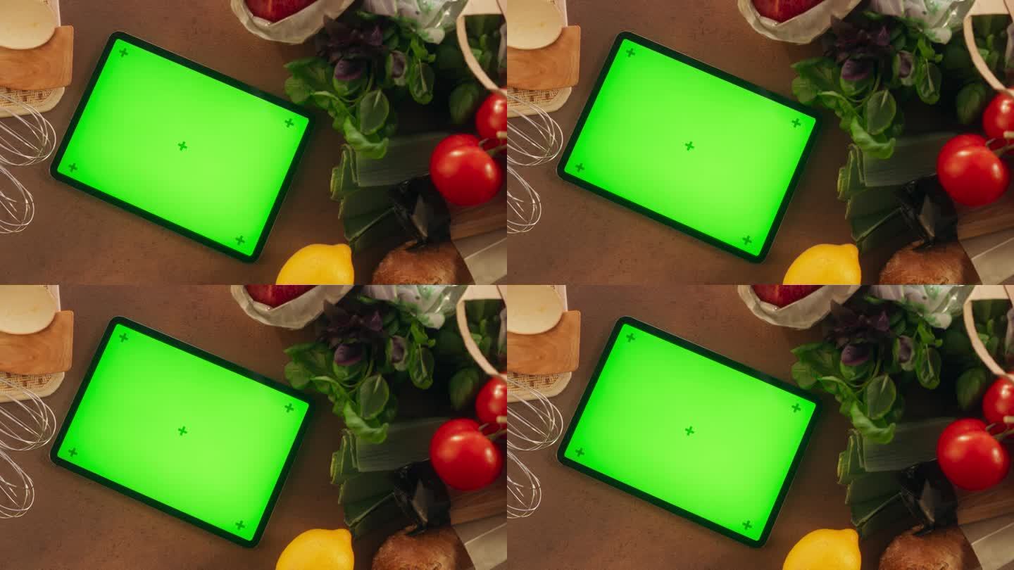 平板电脑的顶部静态视图与模拟绿屏显示。一个设备躺在厨房桌子上，周围都是蔬菜。模板在线数字营销的食品商