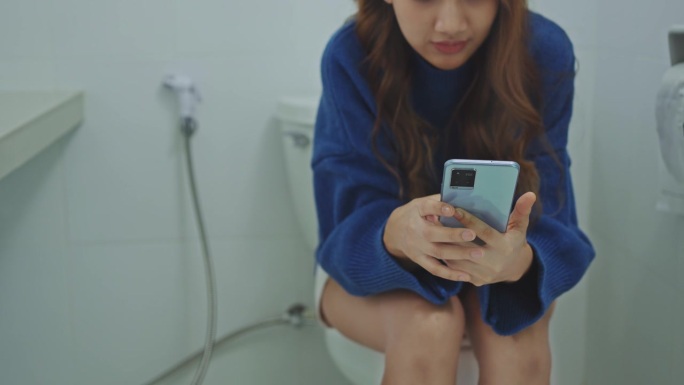 年轻女子坐在家里卫生间的马桶上使用智能手机。