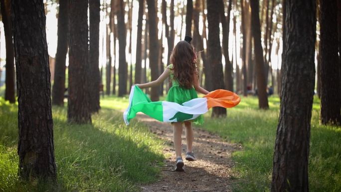 小女孩庆祝圣帕特里克节。可爱的9岁孩子穿着绿色的小精灵服装，带着爱尔兰国旗慢镜头跑过森林