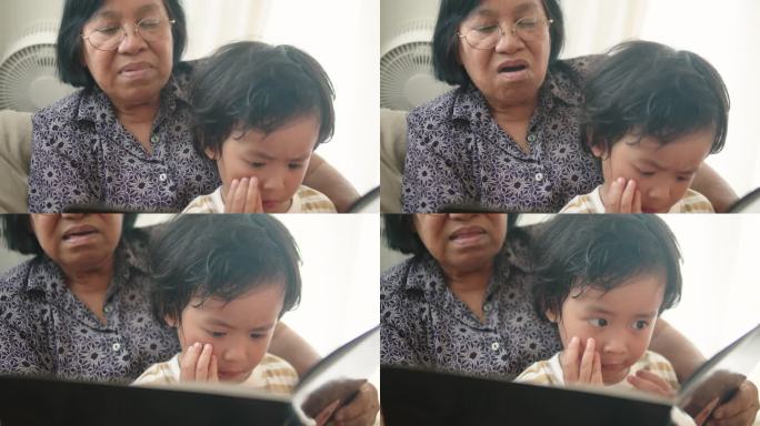 可爱的亚洲孙子和奶奶在沙发上看书。蹒跚学步的男孩和他的奶奶一起学习阅读。
