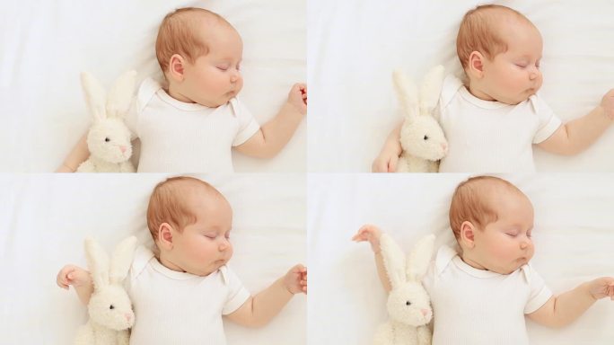 可爱的刚出生的小女孩，手里抱着一只毛绒兔子，在家里的白棉床上甜甜地睡着了，一个安静的小婴儿睡在婴儿床