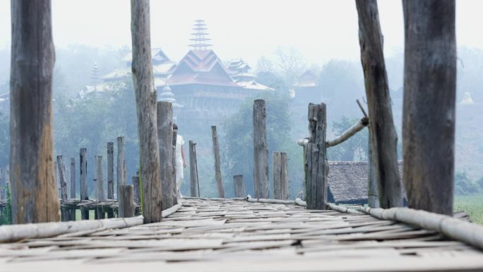 在一个雾蒙蒙的清晨，一位身穿白衣的年轻女士在一座木桥上漫步，向镜头靠近。