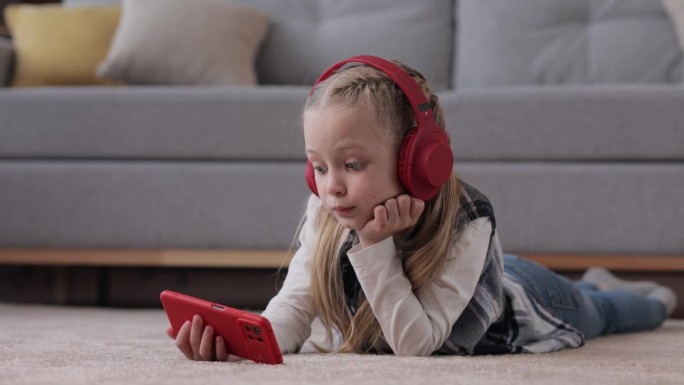 惊讶的小女孩戴着红色耳机躺在家里的地毯上。可爱的女孩拿着智能手机，看动画片，发短信，浏览互联网和社交