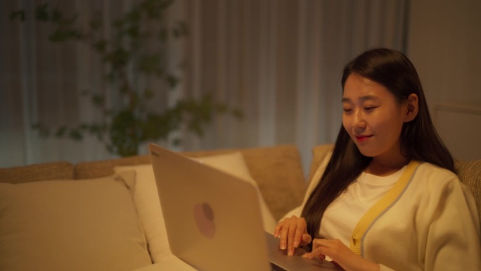 受启发的韩国自由撰稿人在舒适的沙发上放松工作。深夜，在极简主义的客厅里，一位女士在笔记本电脑上写了一