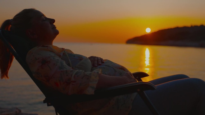微笑的孕妇触摸她的肚子，而放松在甲板上的椅子与美丽的大海和天空的背景在日落的侧视图