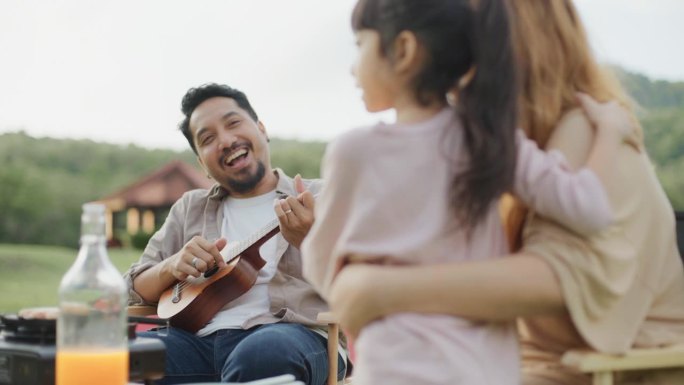 快乐的亚洲家庭在公园野餐。父亲展示唱歌和弹奏尤克里里。