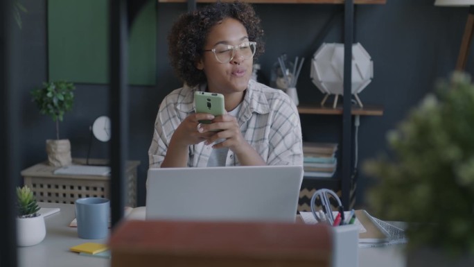 自由职业者的肖像，非洲裔美国人使用智能手机短信，然后在办公桌上用笔记本电脑工作