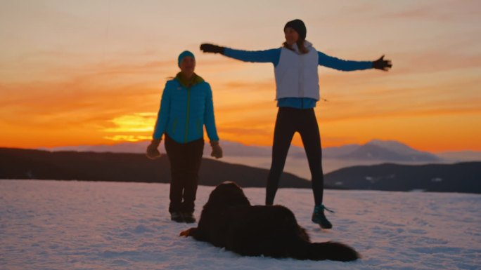 两位女士带着她们的狗在雪地里远足前正在做热身运动