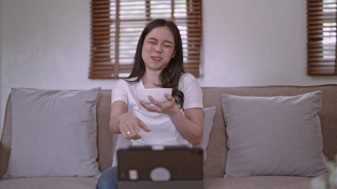 亚洲女性吃零食和看搞笑电影在便携式平板电脑上，不健康的生活方式理念。