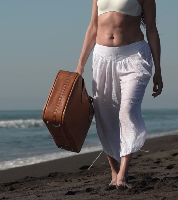 一个成熟的女人带着棕色的手提箱走在太平洋的黑色沙滩上