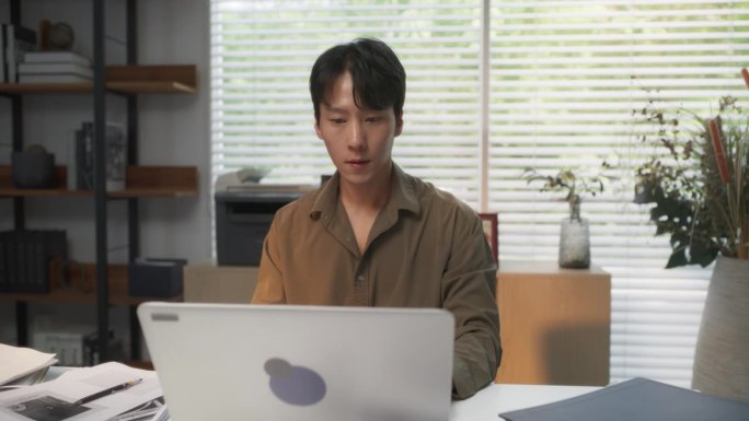 年轻英俊的韩国专业人士，在他舒适的家庭办公室里，沉浸在笔记本电脑的办公桌后面，用建筑的眼光制作现代设