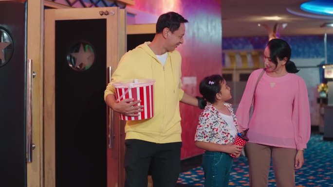 快乐的亚洲家庭在电影院门口吃着爆米花和饮料。一对父母带着女儿看完电影走出了电影院的出口。快乐亚洲家庭