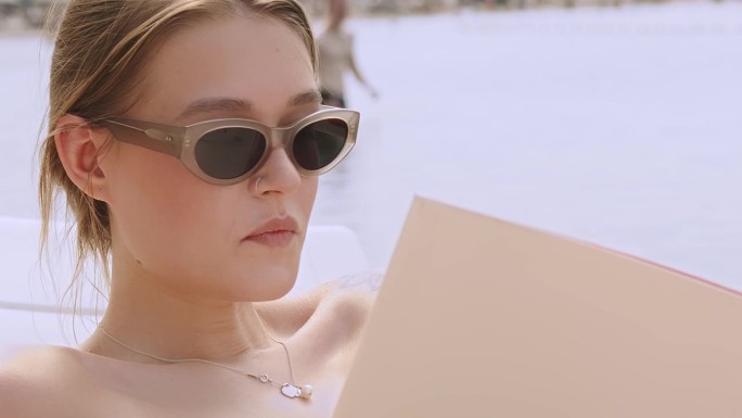 一个戴着墨镜的女孩在沙滩上的画像。一个年轻的美女躺在沙滩上或泳池边读杂志或书。在背景中，一个男孩和一