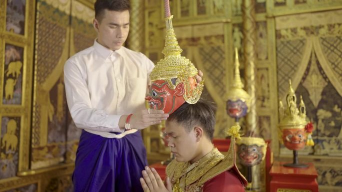一名身穿泰式布料的亚洲男子手持孔庙面具，站在那里进行传统的敬师仪式。