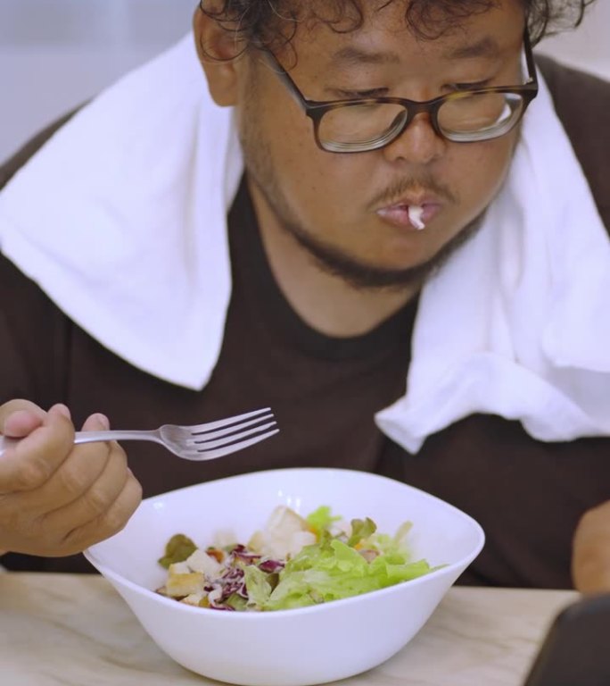 痴迷的亚洲男子一边吃沙拉一边在手机上看视频，想着自己的减肥目标