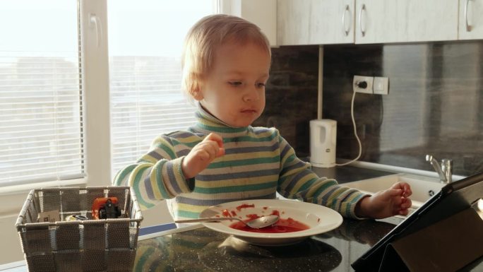 小男孩一边喝汤一边用平板电脑看视频。家庭食品，小玩意上瘾，孩子健康营养