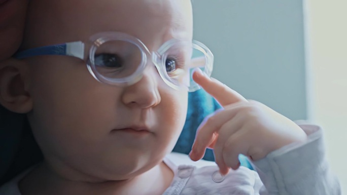 一个一岁的小女孩在看电视的时候用手指碰坏了眼镜。白种人婴儿有斜视、眼疾。视力不佳。孩子坐在妈妈的腿上