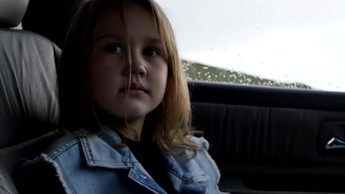 一个可爱的欧洲七岁女孩穿着牛仔背心坐在车里看着。下雨天，一个小女孩坐在一辆有真皮座椅的汽车里的副驾驶