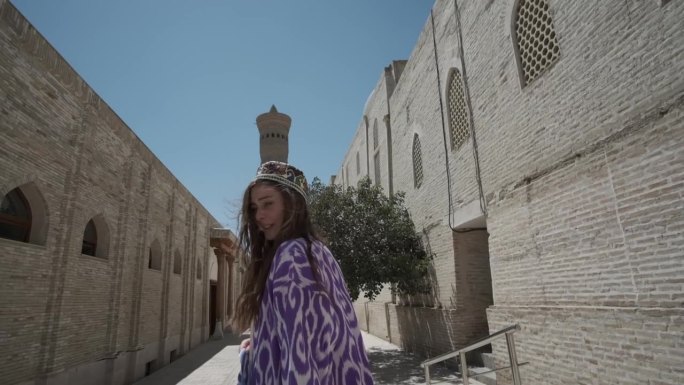 一位身穿东方长袍、头戴无边便帽、长发飘飘的年轻女子站在一座古老的清真寺附近