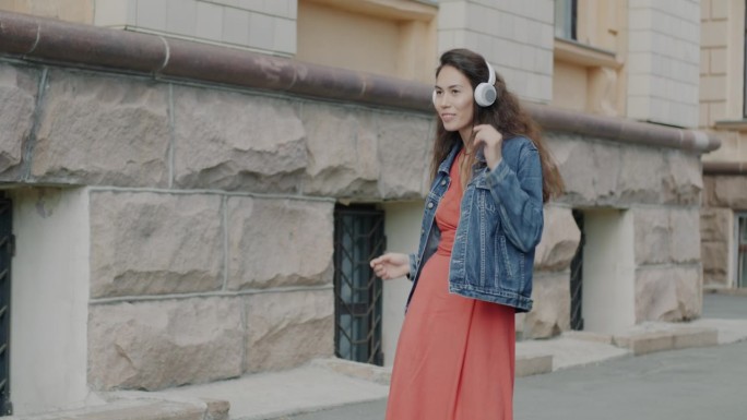美丽的年轻亚洲女子在城市街道上通过耳机听音乐跳舞的慢动作