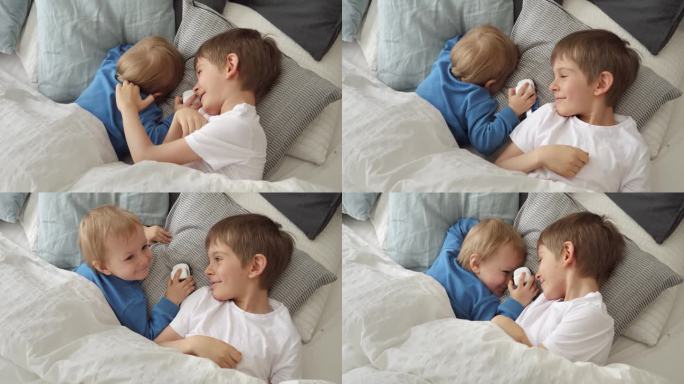 两个可爱的男孩躺在床上柔软的靠垫上，互相微笑。快乐童年的概念，在家里的乐趣和快乐的孩子