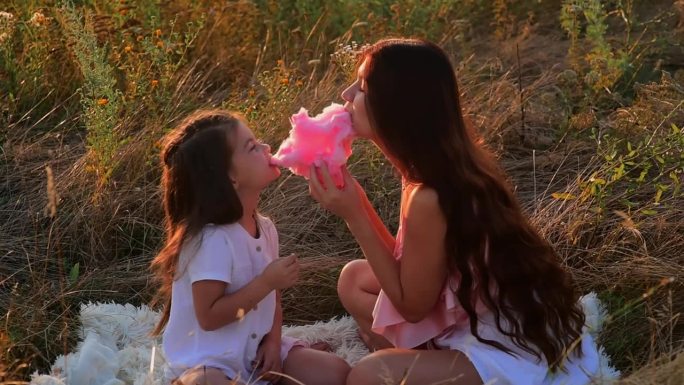 夕阳下，妈妈和女儿在田野里吃粉红色的棉花糖。快乐的童年。度过快乐时光。家庭度假