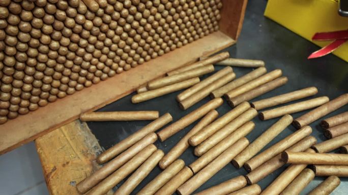 俯视图，许多刚刚卷过的没有标签的手工雪茄被堆放在一堆，随意散落在一张木桌上。这家工厂生产世界上最好的