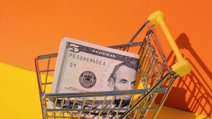 手加5美元钞票在玩具超市购物车黄色背景的钱。销售一篮子美元钞票。最低生活工资概念:贷款、投资养老、储