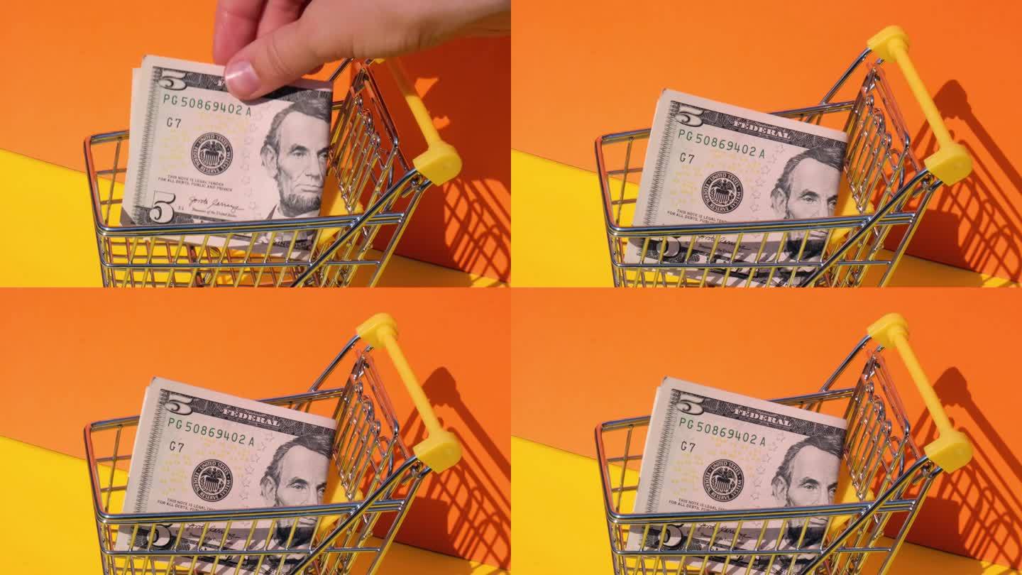 手加5美元钞票在玩具超市购物车黄色背景的钱。销售一篮子美元钞票。最低生活工资概念:贷款、投资养老、储
