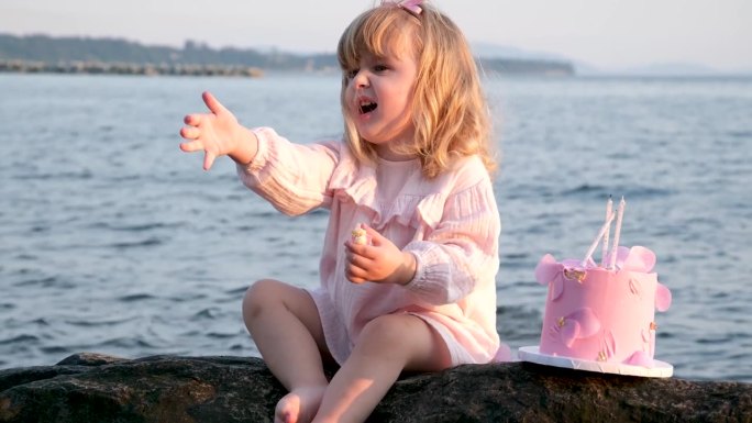 可爱的小女孩，金色的头发，3岁坐在一个蛋糕住妈妈在一个粉红色的裙子，大海坐在一个黑色的石头野餐生日，
