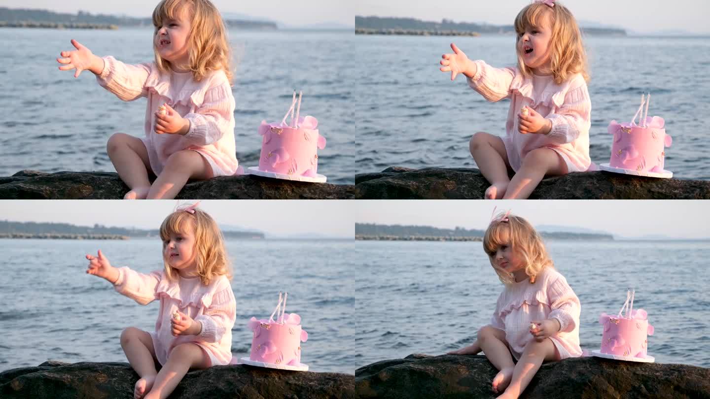 可爱的小女孩，金色的头发，3岁坐在一个蛋糕住妈妈在一个粉红色的裙子，大海坐在一个黑色的石头野餐生日，