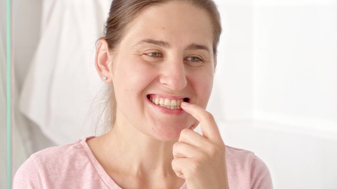 年轻女子在照镜子时用手指擦拭牙菌斑的特写。牙齿健康观念，自我检查口腔及口腔卫生