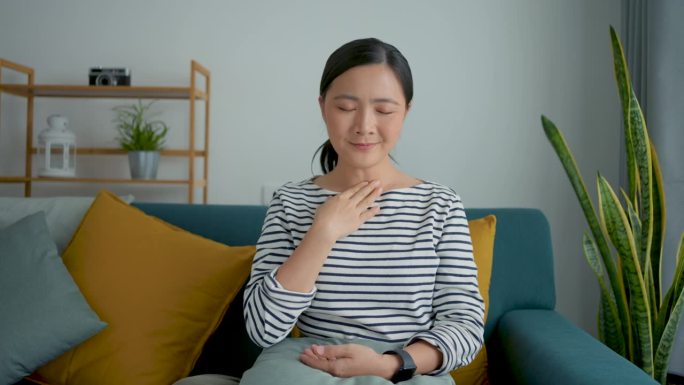 一名亚洲妇女发烧，流鼻涕，鼻塞和喉咙痛，坐在家里客厅的沙发上。