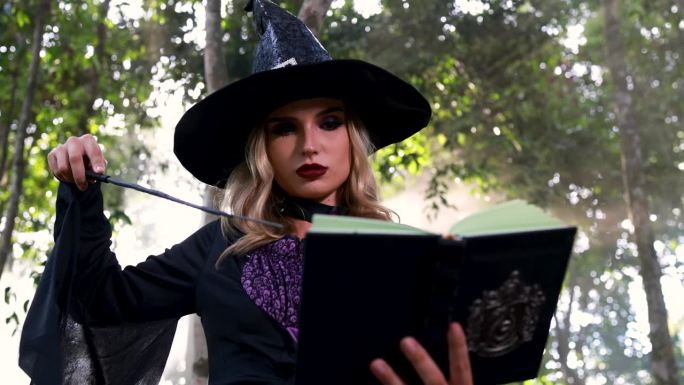 穿着巫婆万圣节服装的美丽女人，手里拿着一本魔法书和一根魔法闪光棒，念着咒语，白色的魔法烟雾从沸腾的罐