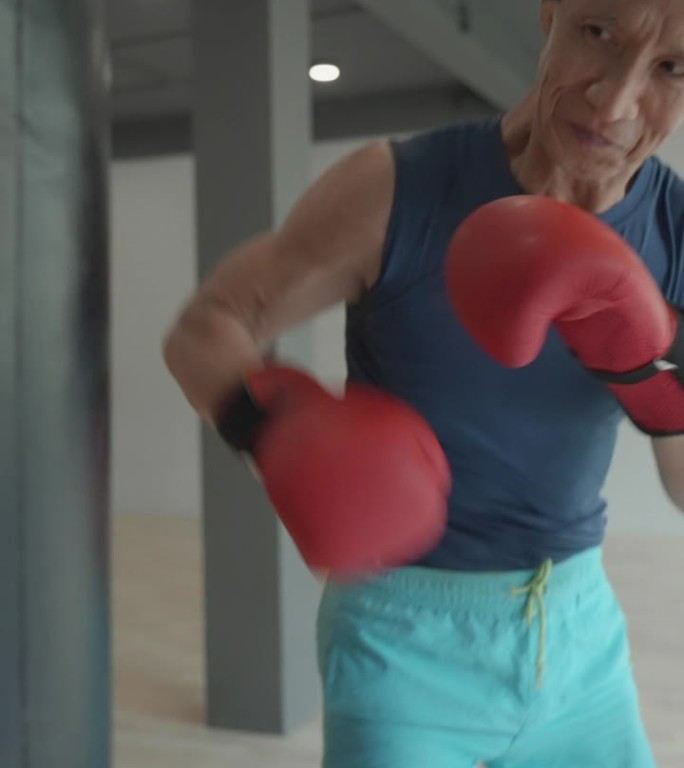 沙袋训练，身穿运动服的亚洲65岁老年肌肉男，在现代健身房拳击沙袋。近距离拍摄一位戴着拳击手套训练的老