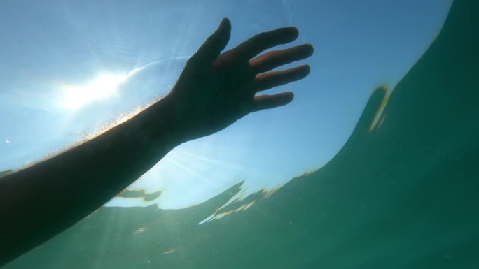 一名溺水者的手漂浮在海水中，背部被夏日炎热的阳光照亮，水下拍摄低角度视角