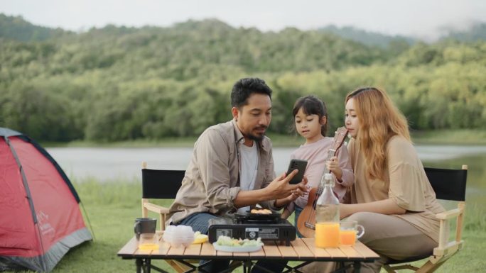 亚洲父母和女儿在阳光明媚的湖边露营地的帐篷里用照相手机自拍。