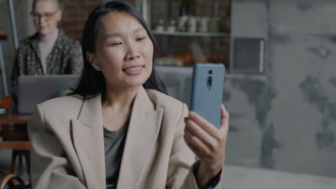 雄心勃勃的亚洲女商人用智能手机和耳机进行在线视频通话，在办公室里挥手致意